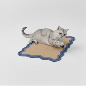 May include: A silver tabby cat rests on a rectangular cat scratcher. The scratcher has a natural tan woven surface with a wavy blue border. The cat is lying down, with its paws extended and its green eyes open.