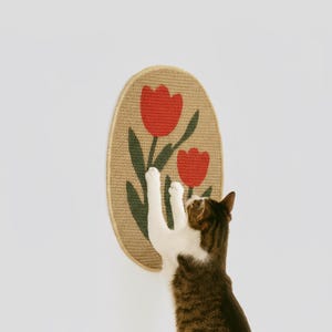 May include: Oval cat scratcher with a woven texture, featuring a red tulip design on a beige background. A brown and white cat is reaching up to scratch the surface. The scratcher is mounted on a white wall.