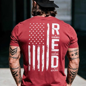 May include: A maroon short-sleeved t-shirt featuring a white distressed American flag graphic on the back. The design includes the vertical text R.E.D. with the words REMEMBER, EVERYONE, DEPLOYED printed below each letter in a white sans-serif font.