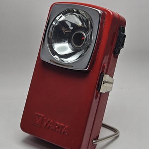 May include: A vintage red Varta torch with a chrome bezel around the lens. The rectangular torch has a black switch and a metal stand. The Varta logo is visible on the front.
