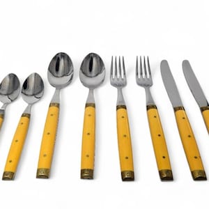 Könnte beinhalten: Ein Set aus Edelstahlbesteck mit gelben Griffen. Das Set enthält Löffel, Gabeln und Messer. Die Griffe sind hellgelb mit messingfarbenen Akzenten. Das Besteck ist auf weißem Hintergrund angeordnet.