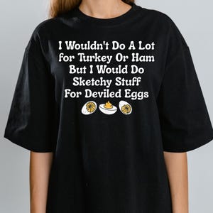 Puede incluir: Camiseta negra con el texto "I Wouldn't Do A Lot for Turkey Or Ham But I Would Do Sketchy Stuff For Deviled Eggs." La camiseta también presenta un gráfico de tres huevos rellenos.