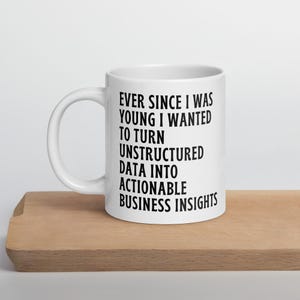 Pode incluir: Caneca de cerâmica branca com o texto "EVER SINCE I WAS YOUNG I WANTED TO TURN UNSTRUCTURED DATA INTO ACTIONABLE BUSINESS INSIGHTS" em preto. A caneca está sobre uma superfície de madeira.