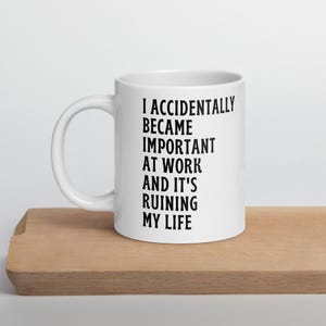 以下が含まれることがあります： 「I ACCIDENTALLY BECAME IMPORTANT AT WORK AND IT'S RUINING MY LIFE」の黒い文字がプリントされた白いセラミックマグ。マグカップは木製の表面に置かれています。マグカップには大きなハンドルが付いています。