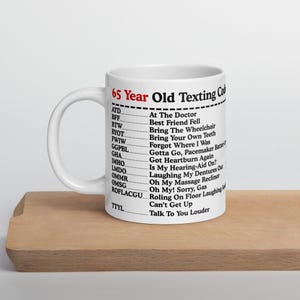 May include: White ceramic mug with the text "65 Year Old Texting Code" and a list of acronyms with their meanings. The mug is on a wooden surface.