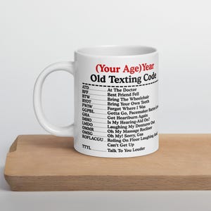 May include: White ceramic mug with the text "(Your Age) Year Old Texting Code" in black and red. The mug lists old texting acronyms and their meanings, such as "ATD" for "At The Doctor".