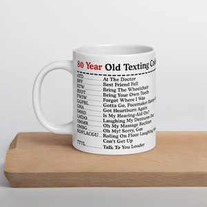 May include: White ceramic mug with the text "80 Year Old Texting Code" and a list of acronyms with their meanings. The mug is on a wooden surface.