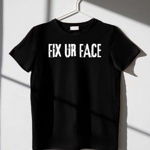 Könnte beinhalten: Schwarzes T-Shirt mit dem weißen Text "FIX UR FACE" in einer verwaschenen Schriftart. Das Shirt hängt an einem weißen Kleiderbügel vor einem weißen Hintergrund. Das kurzärmelige T-Shirt ist aus einem weichen Material.