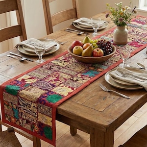 Könnte beinhalten: Ein rustikaler Holztisch, gedeckt für ein Essen. Ein farbenfroher Patchwork-Tischläufer schmückt die Mitte, mit einer Obstschale und einer Blumenvase. Gedecke mit Tellern, Besteck und Servietten sind für die Gäste arrangiert.