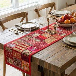 May include: A rustic wooden dining table set for a meal. A red and gold patterned table runner runs down the center, with plates, silverware, and a bowl of fruit. Wooden chairs surround the table.