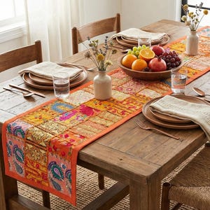 Può includere: Un tavolo da pranzo rustico in legno è apparecchiato per un pasto. Il tavolo è adornato da un runner patchwork colorato, piatti di legno e una ciotola di frutta. Sedie in vimini e vasi con fiori completano l'ambientazione.