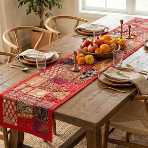 May include: A rustic wooden dining table set for a meal. The table is adorned with a red patterned runner, plates, silverware, and a bowl of fruit. Wooden chairs surround the table, creating a warm and inviting dining space.