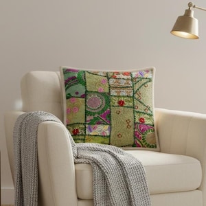 Peut inclure: Un fauteuil blanc cassé avec un coussin patchwork vert, rose et violet. Un plaid en tricot gris est drapé sur le bras. Un lampadaire doré et des plantes en pot complètent la scène du salon.