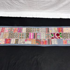 May include: A rectangular patchwork textile with a grey border. The patchwork design features a variety of patterns and colours, including pink, red, grey, and cream. The textile is likely a table runner or decorative piece.