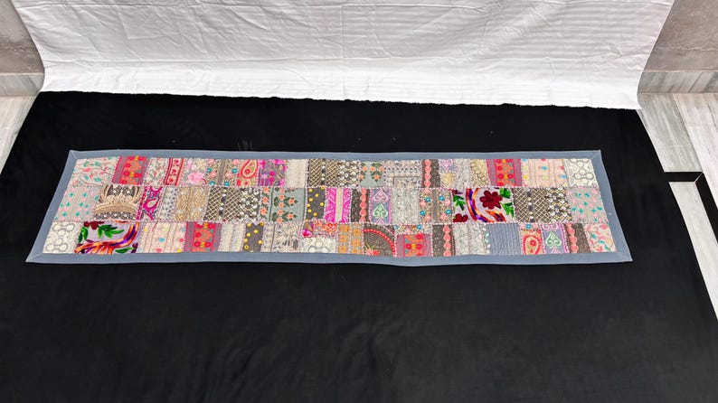 May include: A rectangular patchwork textile with a grey border. The patchwork design features a variety of patterns and colours, including pink, red, grey, and cream. The textile is likely a table runner or decorative piece.