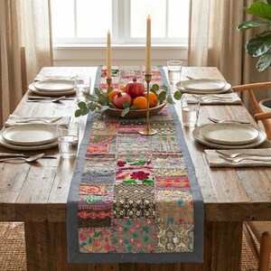May include: A wooden dining table set for a meal, featuring a patchwork runner, plates, cutlery, and candles. A bowl of fruit and greenery decorates the table. The table is set for a gathering.
