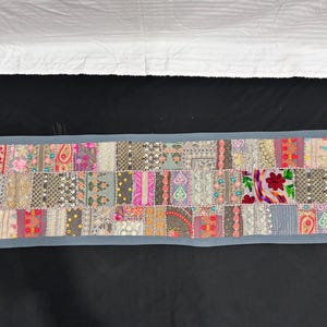 May include: A rectangular patchwork textile with a grey border. The patchwork is made up of various patterned fabric squares in shades of pink, grey, and beige. Floral and paisley designs are visible. The textile is likely a table runner or decorative piece.