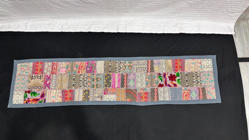 May include: A rectangular patchwork textile with a grey border. The patchwork is made up of various patterned fabric squares in shades of pink, grey, and beige. Floral and paisley designs are visible. The textile is likely a table runner or decorative piece.