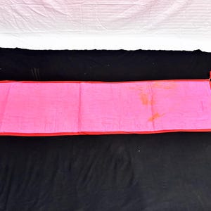May include: A rectangular pink textile with a red border and tassels at each corner. The textile is laid out on a black surface, with a white sheet visible in the background.