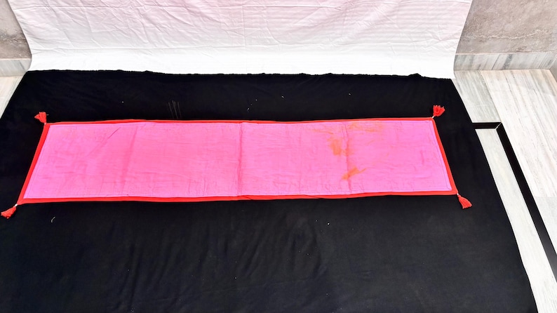 May include: A rectangular pink textile with a red border and tassels at each corner. The textile is laid out on a black surface, with a white sheet visible in the background.