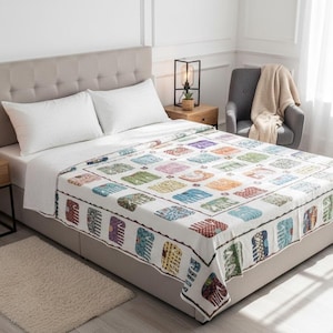 May include: A bed is made with white sheets and pillows, topped with a colorful quilt. The quilt features a repeating pattern of squares in various colors and designs. A neutral rug sits at the foot of the bed, and a chair with a blanket is in the background.