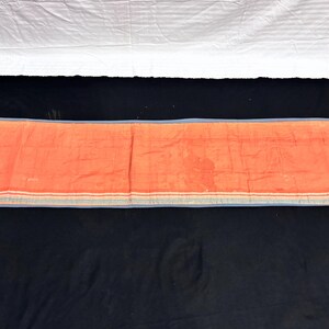 May include: An orange rectangular textile with a blue border and stripes. The textile is laid out on a black surface, with a white sheet visible in the background. The textile appears to be folded.