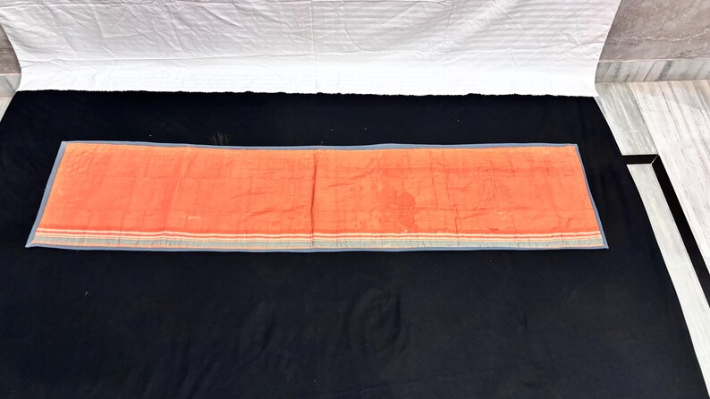 May include: An orange rectangular textile with a blue border and stripes. The textile is laid out on a black surface, with a white sheet visible in the background. The textile appears to be folded.