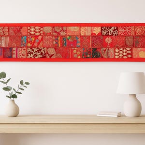May include: A rectangular red wall hanging with a patchwork design. The patchwork is composed of various red fabrics with intricate patterns. The wall hanging is above a wooden table with a vase, plant, lamp, and book.