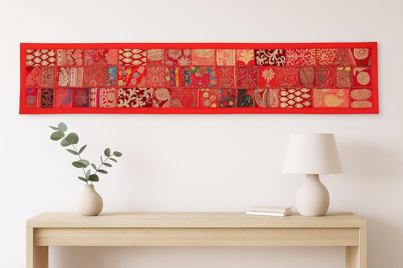 May include: A rectangular red wall hanging with a patchwork design. The patchwork is composed of various red fabrics with intricate patterns. The wall hanging is above a wooden table with a vase, plant, lamp, and book.
