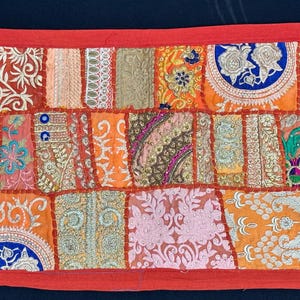 May include: A rectangular patchwork textile with a red border. The design features a variety of embroidered squares in shades of orange, pink, and gold. Intricate floral and geometric patterns adorn the fabric, creating a vibrant and textured appearance.