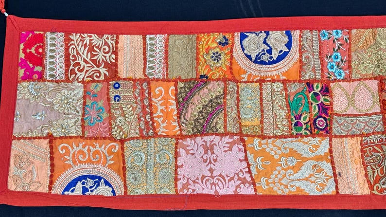 May include: A rectangular patchwork textile with a red border. The design features a variety of embroidered squares in shades of orange, pink, and gold. Intricate floral and geometric patterns adorn the fabric, creating a vibrant and textured appearance.