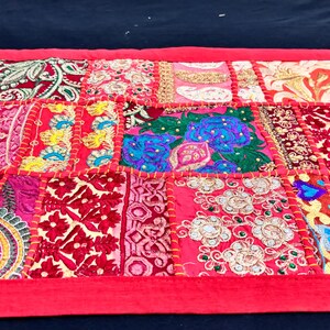 May include: A vibrant, patchwork textile with a red border. The design features a variety of patterned squares in shades of red, pink, and gold, with embroidered details. This decorative piece could be a table runner or wall hanging.