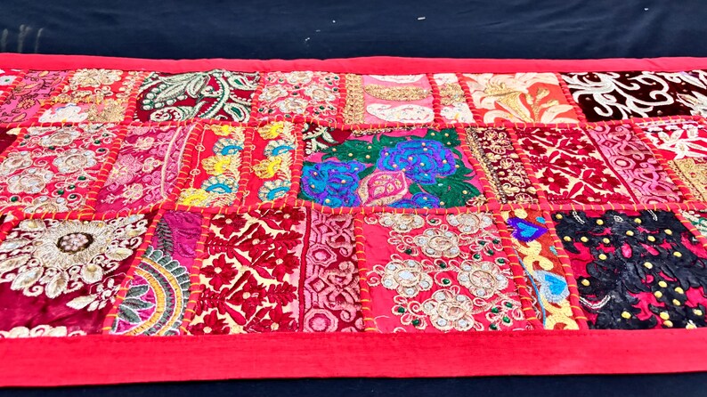 May include: A vibrant, patchwork textile with a red border. The design features a variety of patterned squares in shades of red, pink, and gold, with embroidered details. This decorative piece could be a table runner or wall hanging.