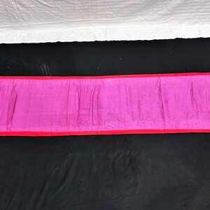 May include: A rectangular, magenta-colored fabric with a red border. The fabric is laid out on a black surface, with a white sheet visible in the background. The item appears to be a decorative textile.