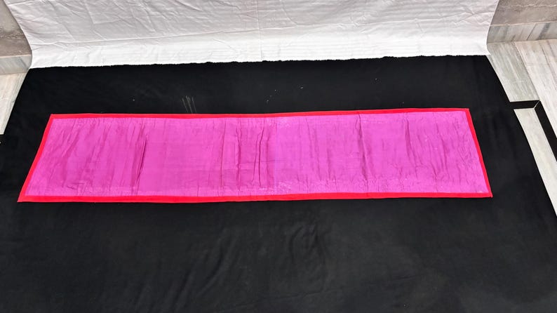 May include: A rectangular, magenta-colored fabric with a red border. The fabric is laid out on a black surface, with a white sheet visible in the background. The item appears to be a decorative textile.