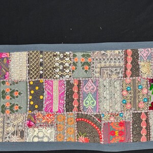 May include: A rectangular patchwork textile with a blue border. The design features a variety of embroidered squares in shades of pink, orange, green, and grey, with floral and geometric patterns. The textile is likely used as a table runner.