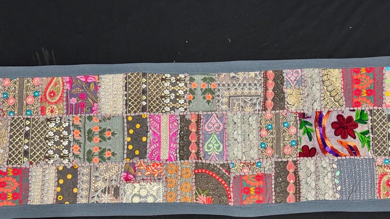 May include: A rectangular patchwork textile with a blue border. The design features a variety of embroidered squares in shades of pink, orange, green, and grey, with floral and geometric patterns. The textile is likely used as a table runner.