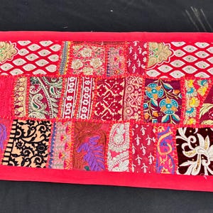 May include: A rectangular patchwork textile in shades of red, pink, and burgundy. The design features a variety of patterns, including paisley, floral, and geometric shapes. The edges are framed with a solid red border. This decorative piece is suitable for home decor.