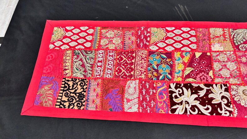May include: A rectangular patchwork textile in shades of red, pink, and burgundy. The design features a variety of patterns, including paisley, floral, and geometric shapes. The edges are framed with a solid red border. This decorative piece is suitable for home decor.