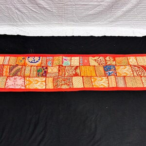 May include: A rectangular, patchwork textile with a red border and tassels at each corner. The patchwork is composed of various fabric squares in shades of orange, yellow, and red, with intricate patterns and designs. The textile is displayed on a black surface.