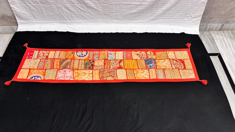 May include: A rectangular, patchwork textile with a red border and tassels at each corner. The patchwork is composed of various fabric squares in shades of orange, yellow, and red, with intricate patterns and designs. The textile is displayed on a black surface.