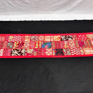 May include: A rectangular, red patchwork textile with a vibrant mix of patterns and colors. The design features a variety of fabric squares in shades of red, pink, and other colors, framed by a solid red border. This decorative piece is likely a table runner.