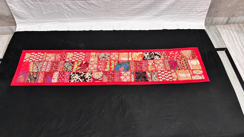 May include: A rectangular, red patchwork textile with a vibrant mix of patterns and colors. The design features a variety of fabric squares in shades of red, pink, and other colors, framed by a solid red border. This decorative piece is likely a table runner.
