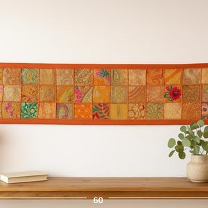May include: A rectangular wall hanging with a patchwork design. The piece features a vibrant mix of orange, yellow, and green fabric squares, framed by a solid orange border. Tassels adorn the corners. The piece is approximately 60 inches wide.