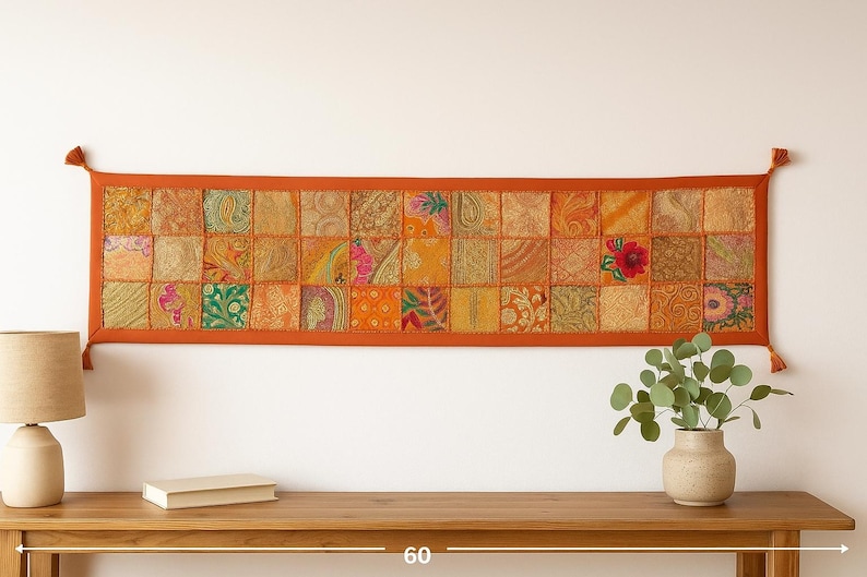 May include: A rectangular wall hanging with a patchwork design. The piece features a vibrant mix of orange, yellow, and green fabric squares, framed by a solid orange border. Tassels adorn the corners. The piece is approximately 60 inches wide.