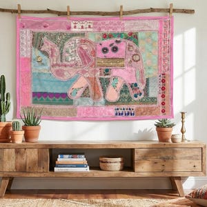 Puede incluir: Un tapiz rosa con un elefante cuelga sobre una consola de madera. El tapiz presenta un diseño de patchwork con un motivo de elefante. Varias plantas en macetas y objetos decorativos están en la consola.