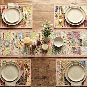 May include: A rustic wooden table set for a meal, featuring four place settings with beige plates, silverware, and patterned placemats. A patchwork table runner with floral and geometric designs runs down the center, with candles and a small vase of flowers.