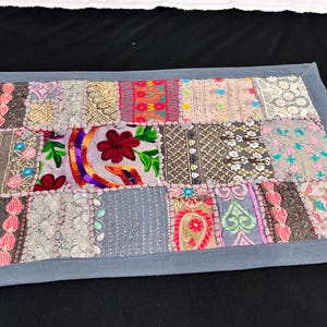 May include: A rectangular patchwork textile with a grey border. The patchwork features various fabric squares in shades of grey, pink, red, and green, with embroidered floral and paisley designs. It is likely a table runner or decorative piece.