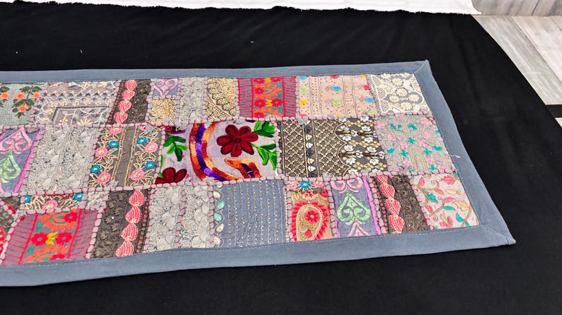 May include: A rectangular patchwork textile with a grey border. The patchwork features various fabric squares in shades of grey, pink, red, and green, with embroidered floral and paisley designs. It is likely a table runner or decorative piece.