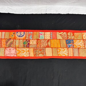 May include: A rectangular, patchwork textile in shades of orange, red, and gold. The design features a variety of patterned fabric squares, framed by a red border. Small red tassels are at each corner. This decorative piece is likely a table runner.